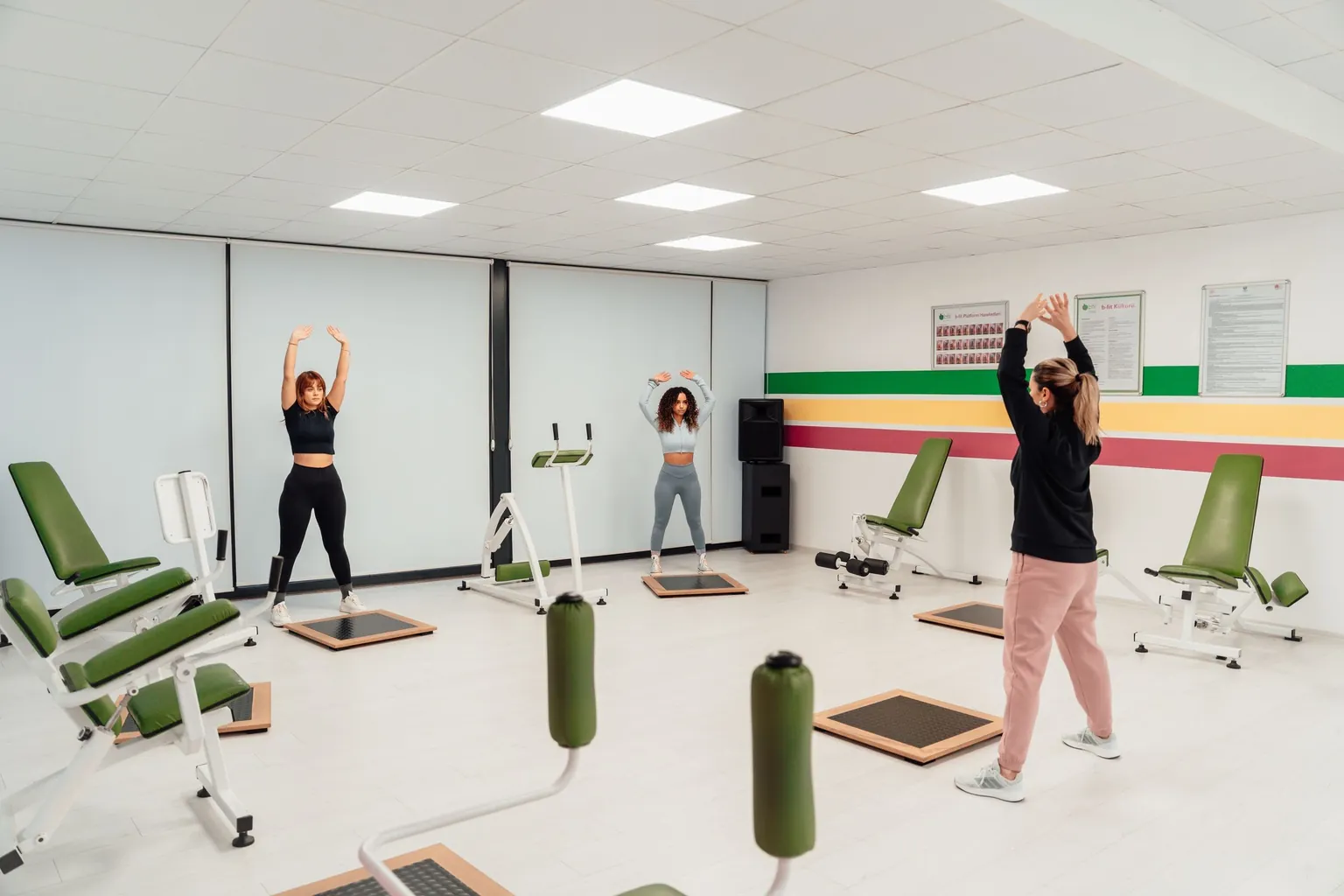 Trois femmes qui s'échauffent bras levés dans une salle équipée de machines de musculation