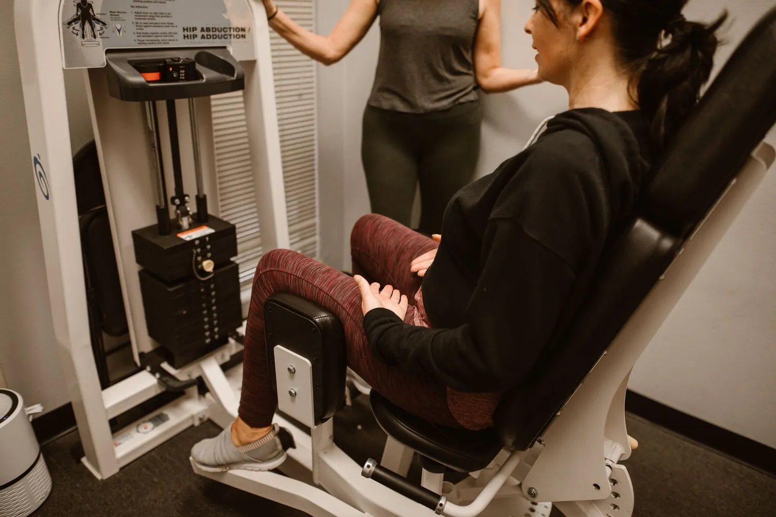 Femme assise sur une machine abduction-adduction, genoux contre les coussinets, une autre personne debout à côté