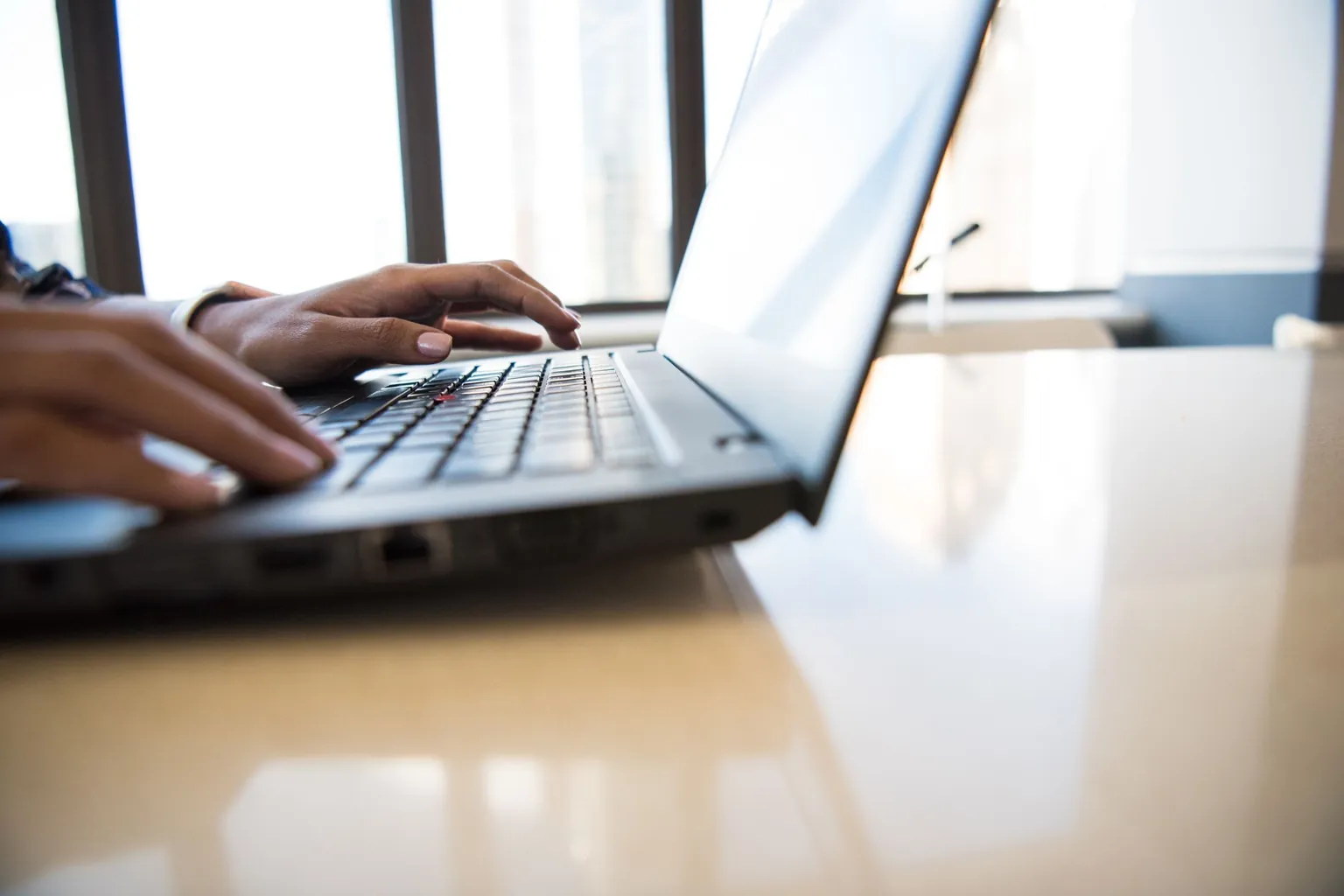 Mains qui tapent sur le clavier d'un ordinateur portable posé sur une table, devant une baie vitrée