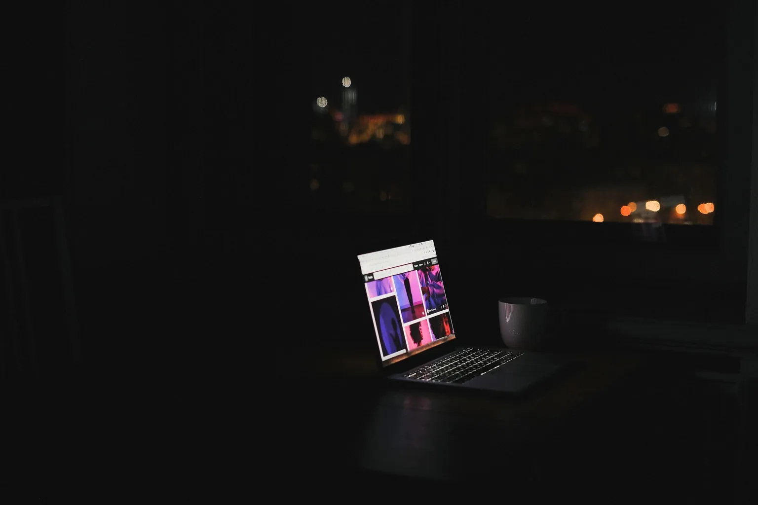 Ordinateur portable allumé sur une table dans une pièce sombre, lumières de la ville derrière la fenêtre