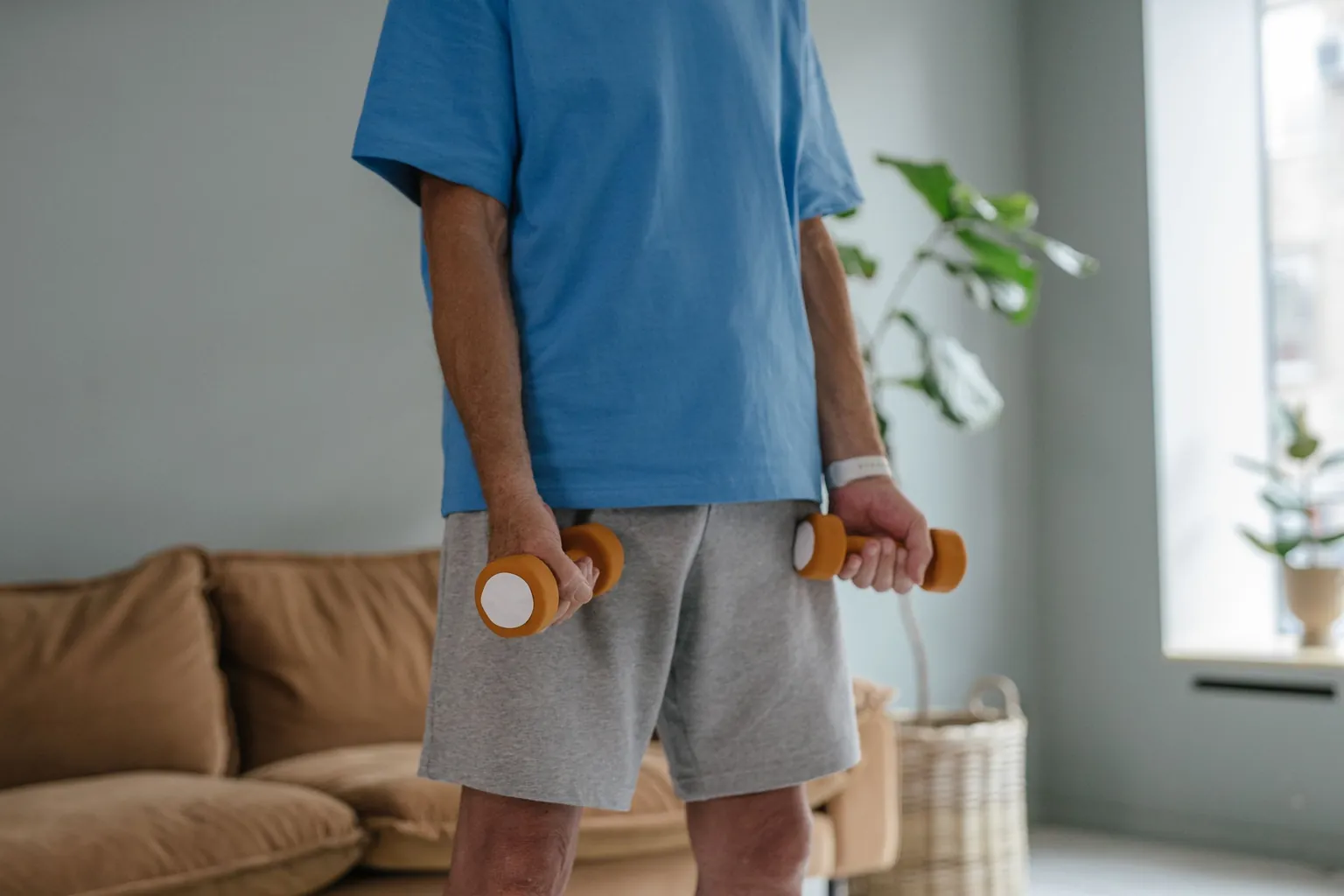 Homme debout dans un salon, un petit haltère orange dans chaque main le long des cuisses