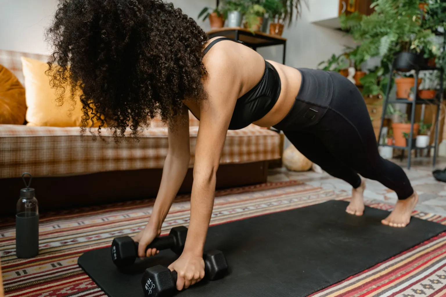 Femme en appui sur deux haltères posés sur un tapis, dans un salon avec canapé et plantes