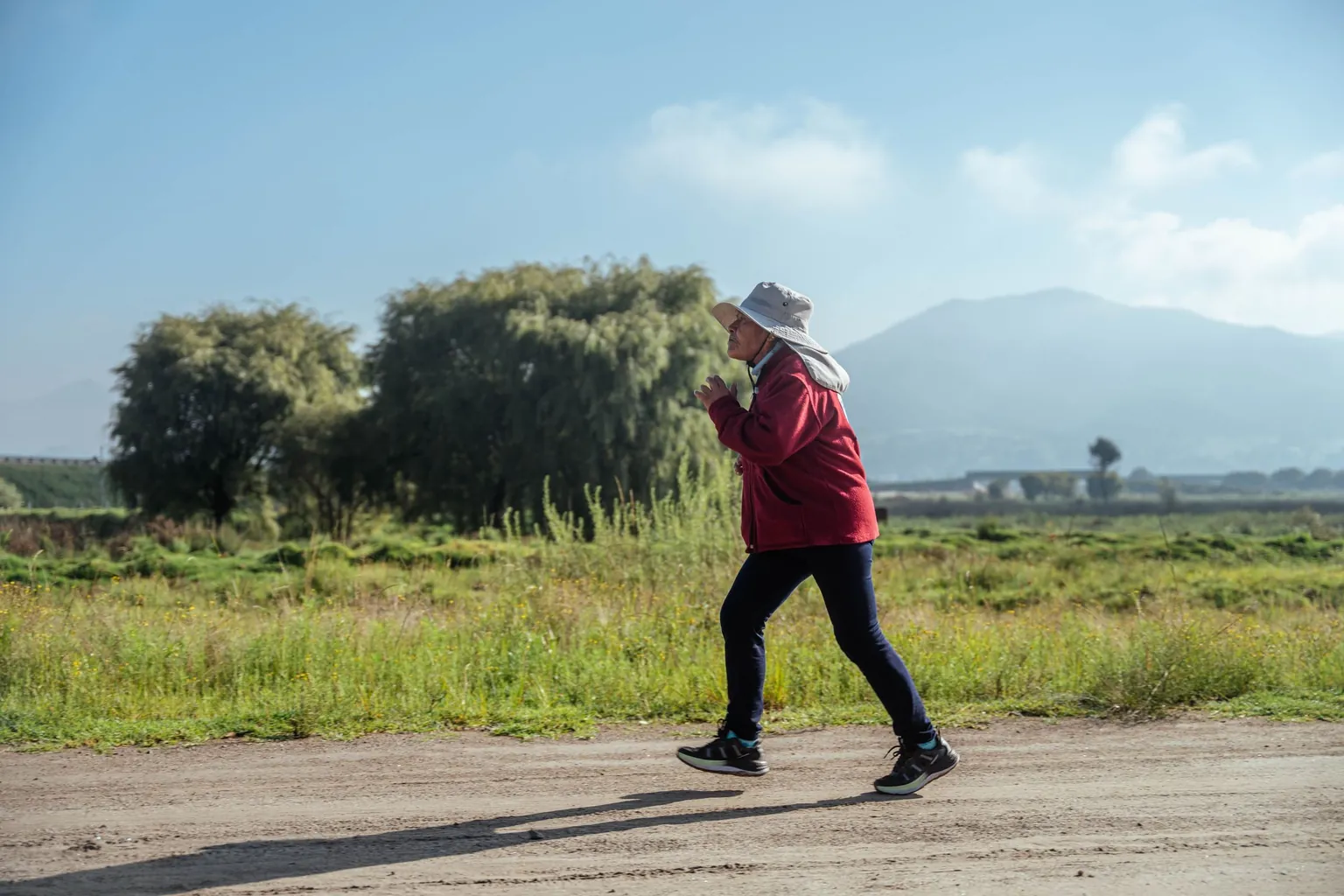 Femme en veste rouge et chapeau qui court à petite allure sur un chemin de terre, à la campagne