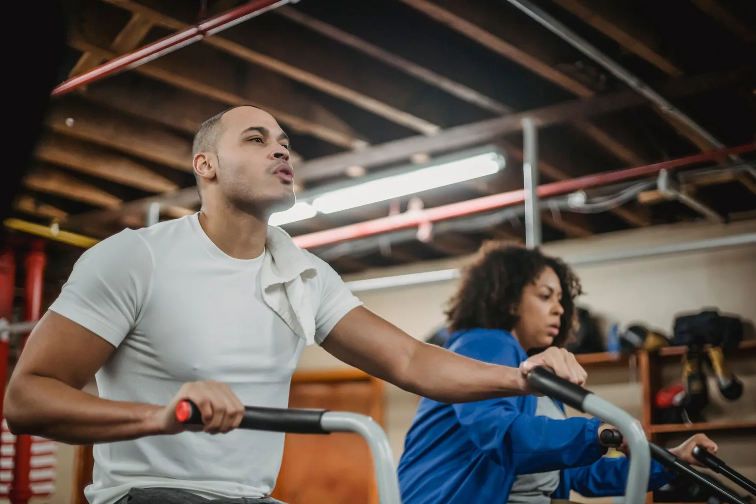 Homme en tee-shirt blanc et femme en veste bleue pédalant sur des vélos à air dans une salle d’entraînement