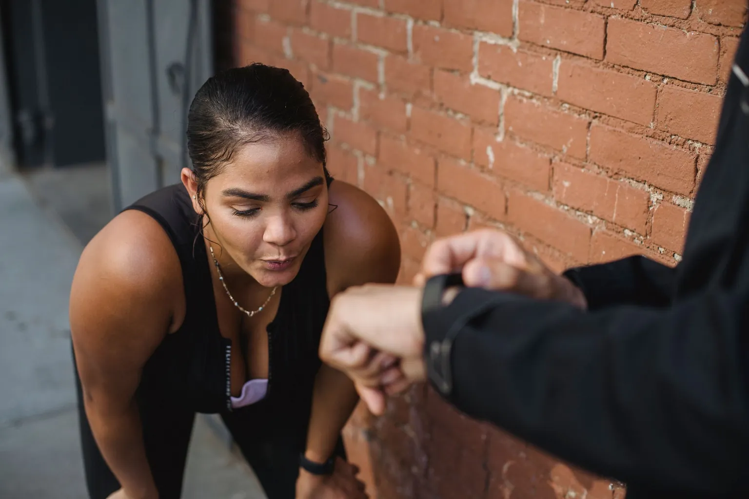 Sportive en débardeur noir, penchée en avant pour reprendre son souffle devant un mur de briques, pendant qu’un partenaire consulte sa montre