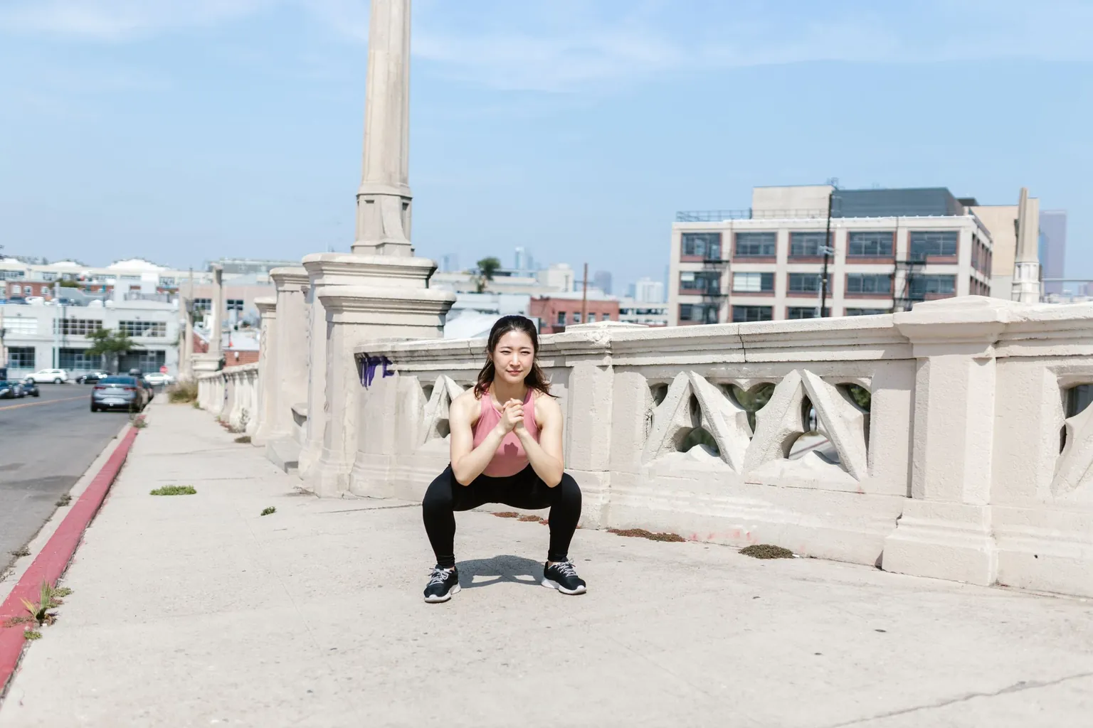 Femme en débardeur rose et legging noir en position de squat pieds écartés sur un trottoir en ville