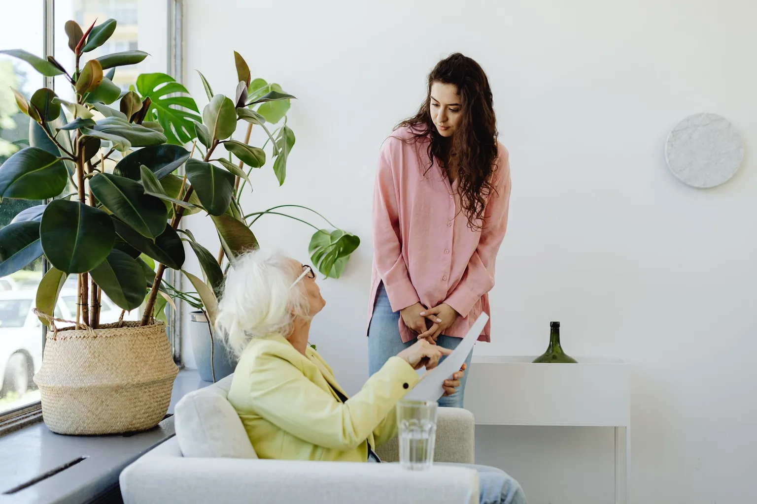 Une femme âgée assise dans un fauteuil montre un document à une jeune femme debout près d'elle, dans un salon lumineux