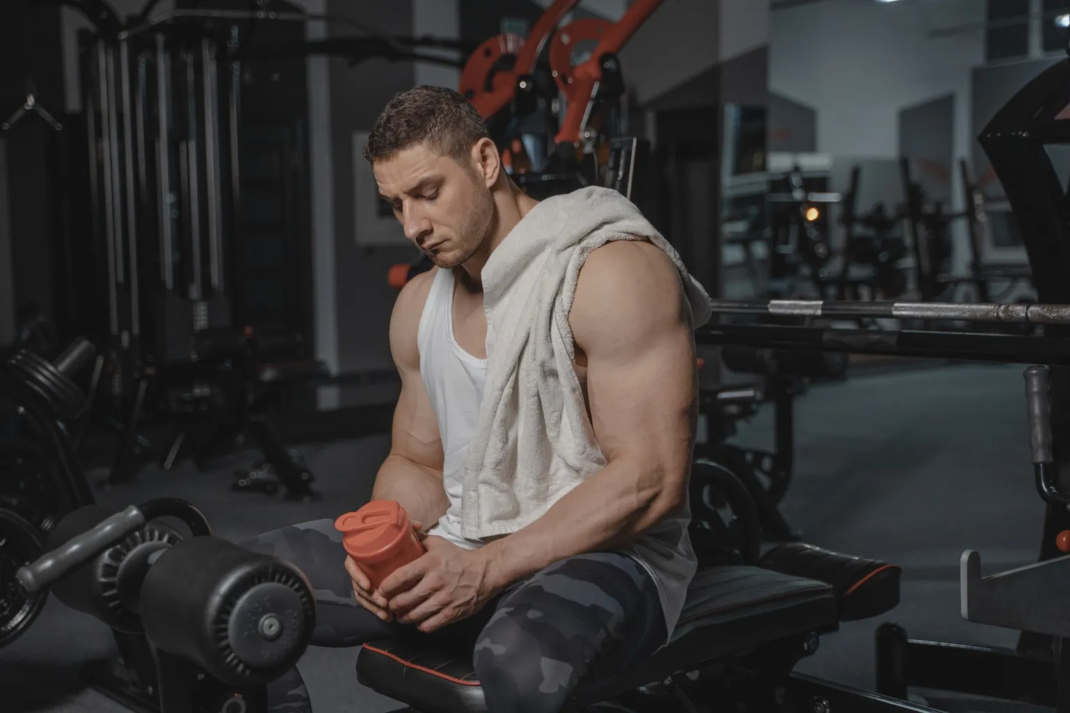 Homme assis sur un banc de musculation, serviette sur l'épaule, tenant un shaker orange