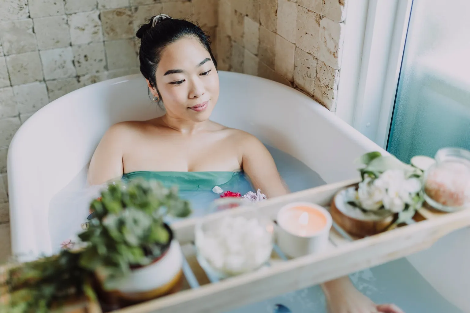 Femme détendue dans une baignoire, plateau avec bougie, fleurs et bocal de sels de bain au premier plan